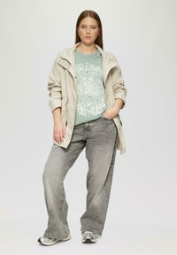 Lichtbeige jack, mintgroene graphic t-shirt met bloemenontwerp, en brede grijze jeans. Zilveren sneakers maken de outfit af.