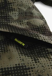 Tarnjacke aus strukturiertem Stoff mit kreisförmigen und sternenförmigen Mustern in Grüntönen und Schwarz, inklusive eines schwarzen Reißverschlussanhängers mit "N2075."