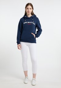 Felpa con cappuccio blu navy con logo "DREIMASTER", tasca a marsupio e cordini bianchi; abbinata a leggings corti bianchi e sneaker.