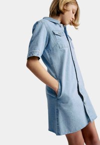 Hellblaues Jeanshemdkleid mit kurzen Ärmeln, Frontknopfverschluss, zwei Brusttaschen und Seitentaschen. Leicht abgerundeter Saum.