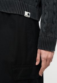 Χέρι που ξεκουράζεται δίπλα στην τσέπη μαύρου παντελονιού εργασίας και σκούρο γκρι πλεκτή ζακέτα με το λογότυπο Carhartt στη ριμπ τελείωμα.