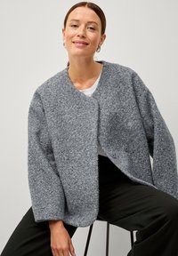 Cardigan grigio con texture, dal taglio morbido, maniche oversize, scollo rotondo e due bottoni sul davanti. Indossato sopra una camicia bianca.