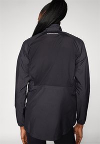 Veste noire légère dotée d'un col montant, de poignets élastiques et d'une taille cintrée. Logo visible sur le haut du dos. Texture lisse.