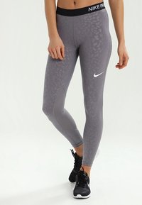 Szare legginsy Nike Pro z teksturowanym, wzorzystym deseniem. Posiadają czarną talię z logo i dopasowany krój. Noszone z czarnymi butami.