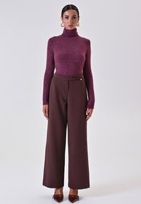 Maglione a collo alto bordeaux con una texture morbida e luccicante abbinato a pantaloni larghi marroni con dettaglio dei bottoni laterali, indossato con scarpe lucide.