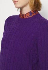 Lila kabelstickad tröja med rund halsringning, över en skjorta i orange och marinblått mönster med vanlig krage.