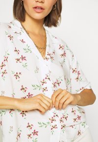 Femme aux cheveux bruns courts portant une chemise blanche à fleurs avec des boutons et des colliers dorés superposés, en train de fermer un bouton à l'avant.