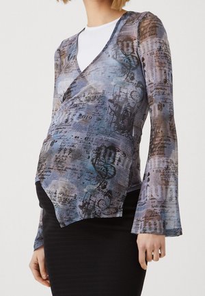 Personne enceinte portant un haut cache-cœur transparent à motifs par-dessus une chemise blanche et une jupe noire texturée, debout devant un fond uni.