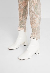Botas de tobillo blancas de charol con punta cuadrada y tacón bloque. Presentan un patrón texturizado de cocodrilo y detalles de tachuelas en el puño.