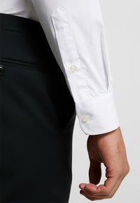 Camisa blanca de vestir con puño puntiagudo, que presenta dos botones y una textura suave; combinada con pantalones negros. Diseño claro y minimalista.