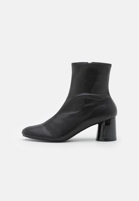 Bottines en cuir noir avec fermeture éclair latérale et un talon en bloc brillant de hauteur moyenne sur fond blanc.