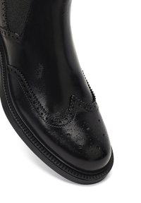 Bota de cuero negro con detalles decorativos de brogue, acabado liso, punta redondeada, paneles elásticos en los costados y suela de goma texturizada.
