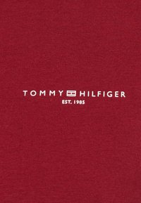 Rødt bomuldsstof med en ribbet tekstur, der har teksten "TOMMY HILFIGER EST. 1985" i hvid, centreret nær bunden.