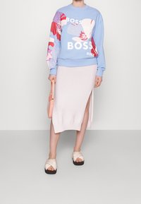 Sudadera azul claro con estampados florales y texto blanco, combinada con una falda rosa claro a la altura de la rodilla con una abertura lateral. Sandalias beige.