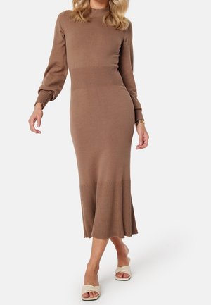 Femme portant une robe midi en maille côtelée marron à manches longues avec un col haut, associée à des chaussures beige ouvertes à enfiler.