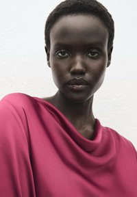 Blouse fuchsia drapée avec une texture douce et un col en cowl, associée à une peau lisse et des cheveux courts sur un fond clair.