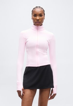 Žena v svetloružovom dlhom tričku na zips s otvorom na palce a čiernej mini sukni, stojaca pred jednoliatym pozadím.