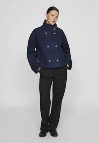 Marineblå ull peacoat med høy krage, dobbelspent design, metallknapper og vide ermer, iført over svarte bukser.