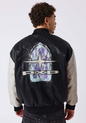 Homme aux cheveux bouclés portant une veste bicolore noire et crème avec un motif floral et étoilé et le texte « Reach for the Stars » dans le dos