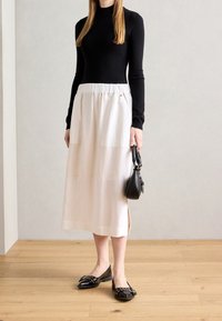 Donna che indossa un top lungo nero, una gonna midi bianca e delle scarpe flat nere con borchie, che tiene in mano una piccola borsa nera, in piedi su un pavimento di legno.