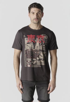 Hombre con camiseta gráfica negra con vistas urbanas de Tokio y Okinawa en rojo y beige, acompañado de pantalones cortos de mezclilla oscuros enrollados.