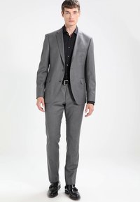 Νεαρός άνδρας στέκεται φορώντας ένα εφαρμοστό γκρίζο κοστούμι με μαύρο πουκάμισο, μαύρη ζώνη και μαύρες επίσημες παπούτσια, απέναντι από έναν απλό λευκό φόντο.