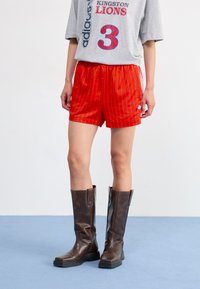 Rode streepjesshorts met een elastische tailleband, gecombineerd met een grijze graphic t-shirt met rode en blauwe tekst. Gedragen met een paar bruine kniehoge laarzen.