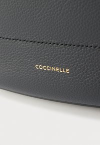 Μαύρη δερμάτινη τσάντα με υφή στην επιφάνεια, που διαθέτει εμφανές ράψιμο και χρυσό ανάγλυφο λογότυπο με την επιγραφή "COCCINELLE".