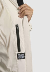Giacca beige chiaro con tasca laterale con zip, accenti neri e una patch testurizzata con la scritta "Khuijo Original Changing Threads."