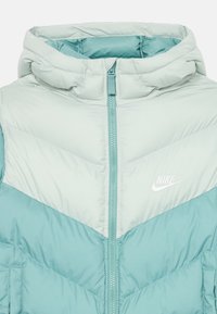 Chaqueta de plumas acolchada ligera en verde menta, con cierre de cremallera, capucha y un diseño en chevron en contraste. Logo de Nike en el pecho.