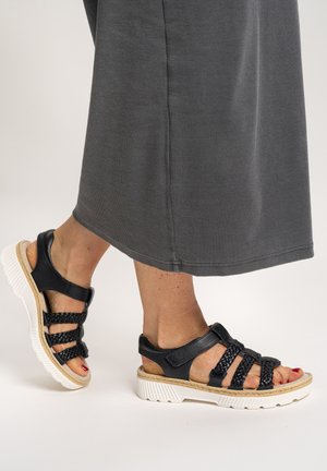 Feet wearing black braided strap sandals with white soles, paired with a gray ribbed long skirt.