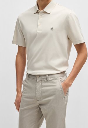 Mann trägt cremefarbenes kurzärmliges Poloshirt mit Knöpfen und hellbeige Hose, eine Hand in der Tasche vor neutralem Hintergrund.