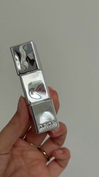 Mano che tiene un astuccio metallicoh a forma quadrata per rossetto con la scritta "TURTLE" in rilievo sul quadrato inferiore, su uno sfondo semplice.