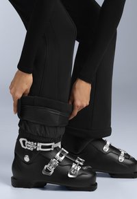 Osoba obutá v černých lyžařských botách se stříbrnými přezkami si upravuje černé lyžařské kalhoty přes elastické návleky, na jednobarevném světle šedém pozadí.