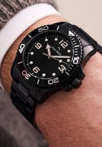 Montre en acier inoxydable noir avec un cadran noir, des index argentés et des chiffres blancs. Elle possède une lunette tournante et un bracelet en maillons métalliques.