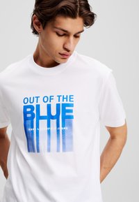 Balta medvilnės marškinėliai su mėlynu gradientiniu užrašu "OUT OF THE BLUE" ir vertikaliomis juostelėmis, o apačioje užrašas "KARL LAGERFELD JEANS". Trumpomis rankovėmis.