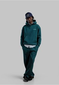 Felpa verde scuro con tasca a marsupio e logo, abbinata a pantaloni coordinati. Sotto, una camicia a strisce blu chiaro e un cappellino blu navy. Scarpe nere.
