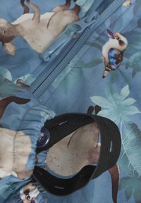 Lätt blått tyg med ett naturinspirerat tryck med djur och bladverk. Inkluderar en blixtlåsstängning och en svart elastisk ögla.