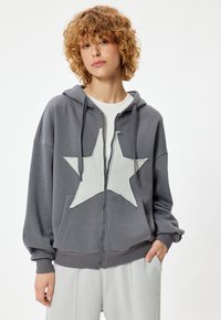 Szara bluza z kapturem na zamek błyskawiczny, z dużym białym wzorem gwiazdy z przodu, oferująca luźny krój, regulowany kaptur oraz miękką, teksturowaną tkaninę.