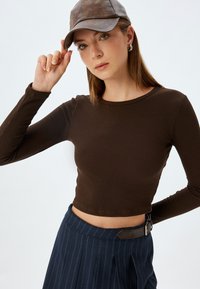 Brązowy, prążkowany, długi rękaw, krótki top, w połączeniu z granatowymi spodniami w paski, o wysokim stanie. Modelka nosi brązową skórzaną czapkę i kolczyki w kształcie kół.