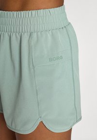 Light green athletic shorts with an elastic waistband, side pocket, and subtle logo detail. Fabric has a smooth texture and scalloped hem.