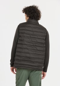 Svart pufferväst med quiltad textur, hög krage, bärs över en långärmad skjorta med gröna cargobyxor synliga. Lättviktig isoleringsdesign.