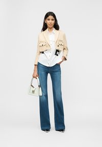 Γυναίκα με κοντό μπεζ σακάκι, λευκό πουκάμισο με κουμπιά, καμπάνα μπλε τζιν, κρατώντας μια μικρή λευκή τσάντα, στέκεται μπροστά σε λευκό φόντο.