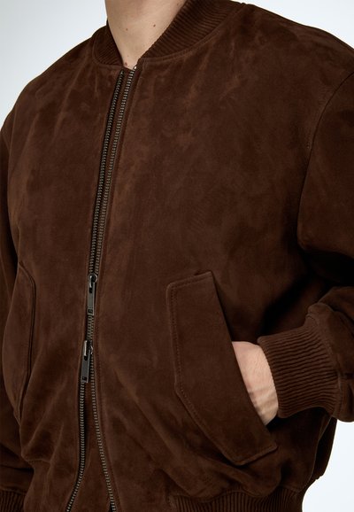 Mann trägt eine dunkelbraune Wildleder-Bomberjacke mit geripptem Kragen und Bündchen, Hand teilweise in der vorderen Tasche, Reißverschluss geschlossen.