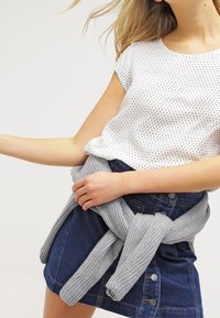 Bílý top s krátkým rukávem a černými puntíky, šedý pletený svetr uvázaný kolem pasu a tmavá džínová sukně s knoflíky vpředu.