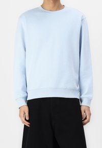 Sweat-shirt bleu clair à col rond en tissu doux, avec des manches longues et des poignets et ourlet côtelés, associé à un pantalon foncé.