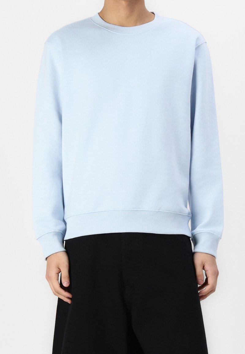 Sweat-shirt bleu clair à col rond en tissu doux, avec des manches longues et des poignets et ourlet côtelés, associé à un pantalon foncé.