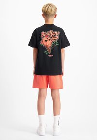 Zwarte short-sleeve t-shirt met een kleurrijke ijscremgraphics en tekst op de achterkant, in combinatie met oranje shorts en witte sneakers.
