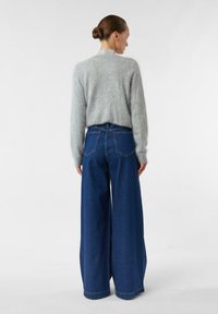 Jasnoszary, puszysty sweter z wysokim kołnierzem, zestawiony z szerokimi, ciemnoniebieskimi jeansami z tylnymi kieszeniami i surowym wykończeniem.