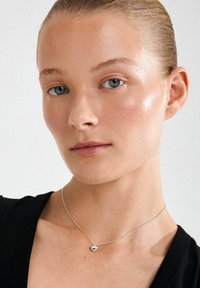 Sidabro širdies formos vėrinys ant plonos grandinėlės, nešiojamas asmens su šviesia oda ir mėlynomis akimis, šviesaus fono priešakyje.